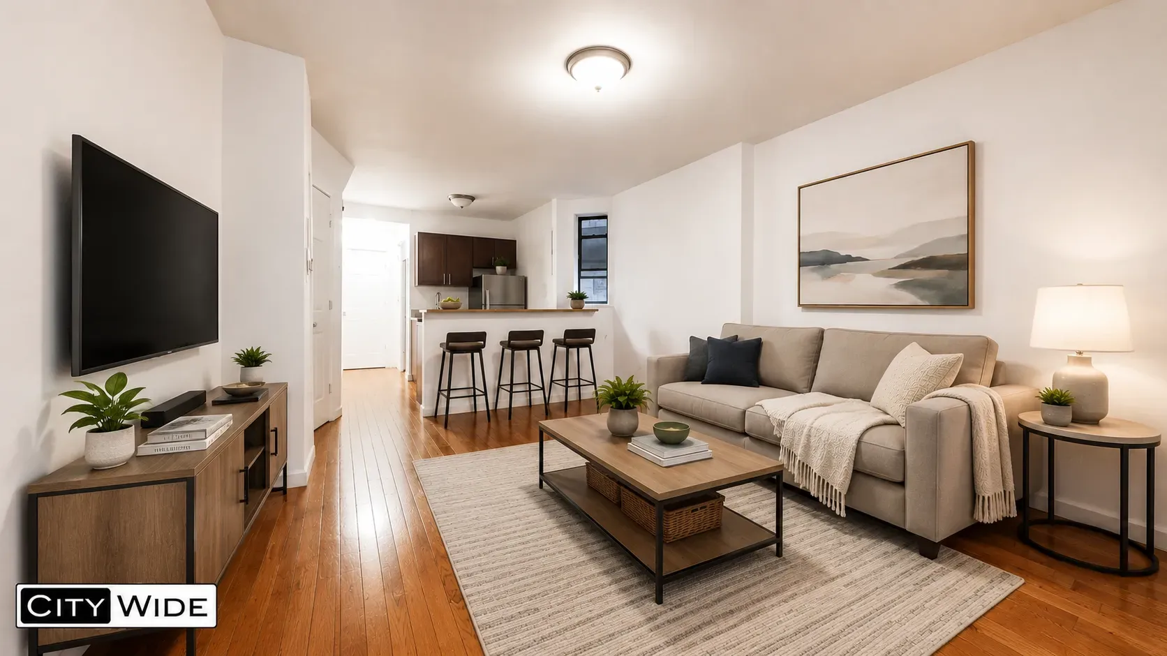 Open-concept living room with hardwood floors, beige sofa, TV, kitchen with bar seating, and framed landscape artwork.