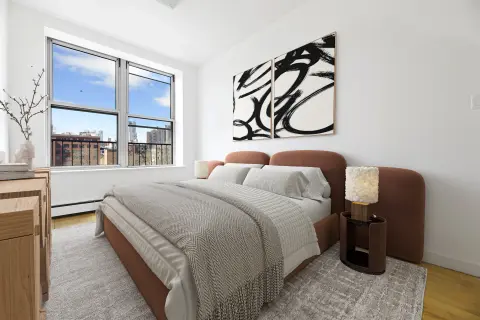 Bedroom with brown bed frame, gray bedding, large window with city view, abstract wall art, and wooden furniture.