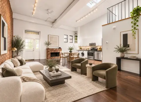 Open-plan loft with exposed brick, cream seating, dark wood floors, kitchen area, and mezzanine level with plants.