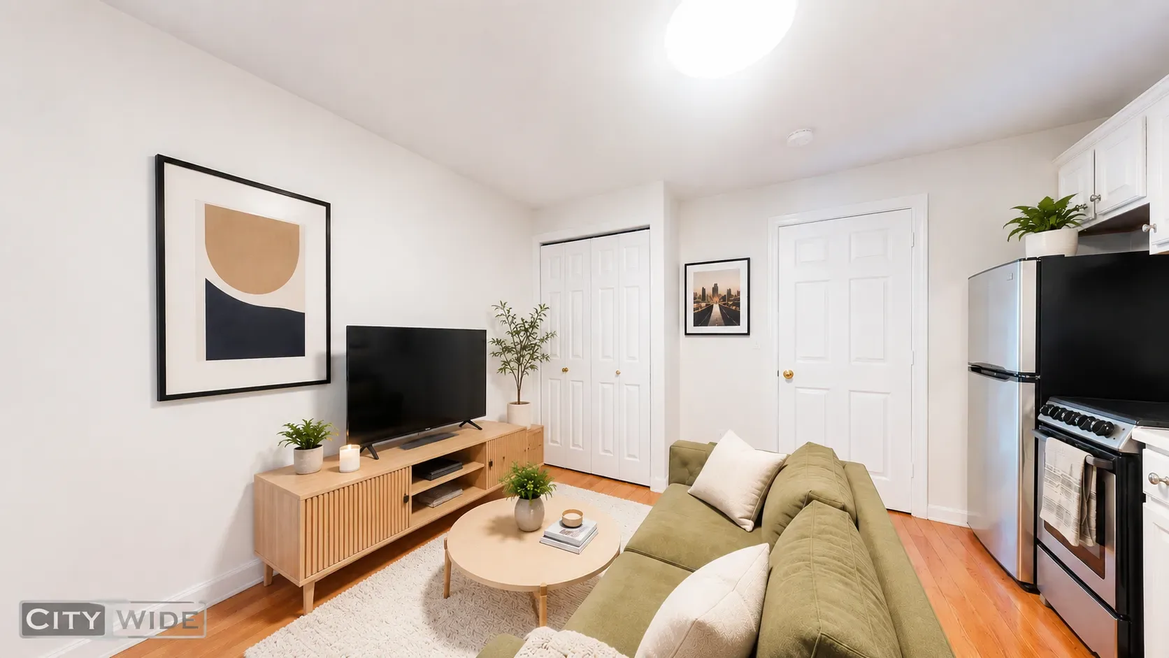 Living room with green sofa, wooden TV stand, framed wall art, white doors, and kitchen area with stainless steel appliances.