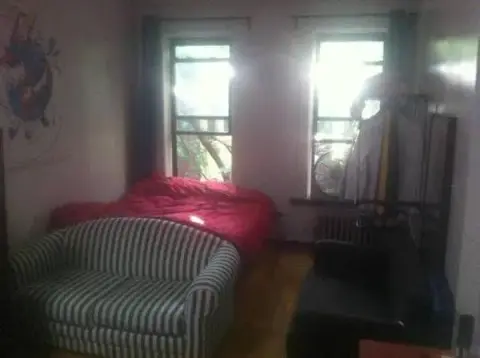 Bedroom with red bed, striped armchair, hardwood floors, two windows with balcony access and radiator.