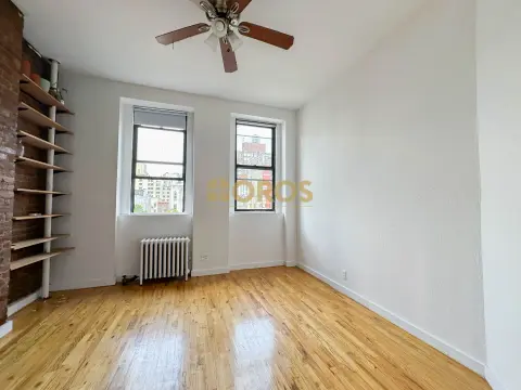 Empty bedroom with hardwood floors, two windows, ceiling fan, radiator, and built-in shelving on one wall.