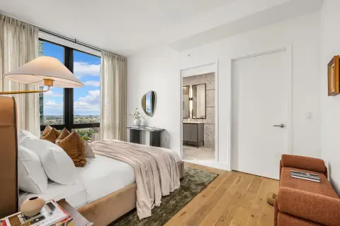 Bedroom with queen bed, large windows with city views, hardwood floors, and ensuite bathroom visible through open doors.