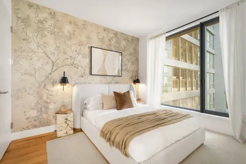 Bedroom with white bed, patterned wallpaper accent wall, pendant lights, large window with city view, and hardwood flooring.