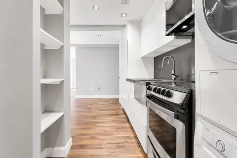 Galley kitchen with white cabinetry, stainless steel appliances, wooden flooring, and open shelving on the left wall.