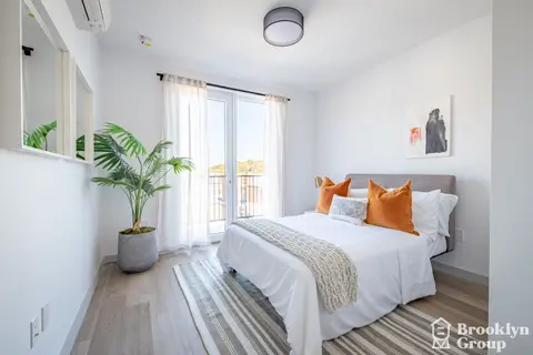 Bedroom with queen bed, orange pillows, potted plant, glass door to balcony, and striped area rug.