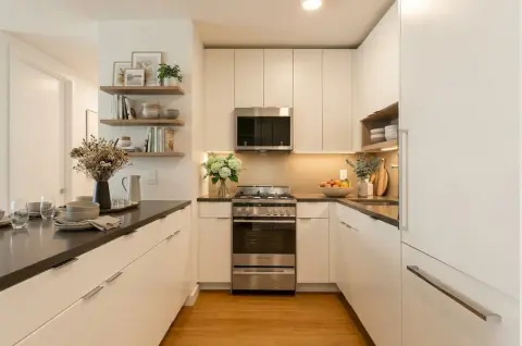 Modern kitchen with white cabinetry, stainless steel appliances, black countertops, open shelving, and wooden flooring.