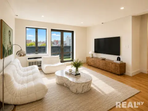 Bright living room with large windows, hardwood floors, white quilted seating, stone coffee table and wooden media console.