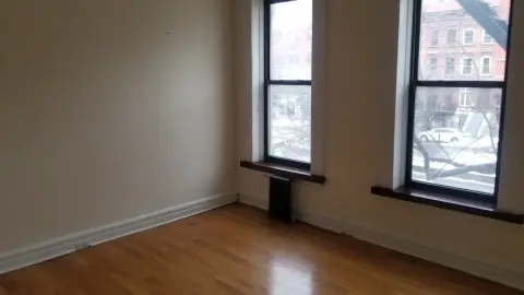 Empty room with hardwood floors, two large windows with black frames, and beige walls.