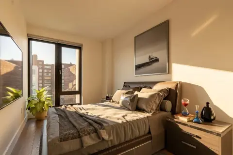 Bedroom with queen bed, wooden nightstand, large windows with city views, and potted plant on windowsill.