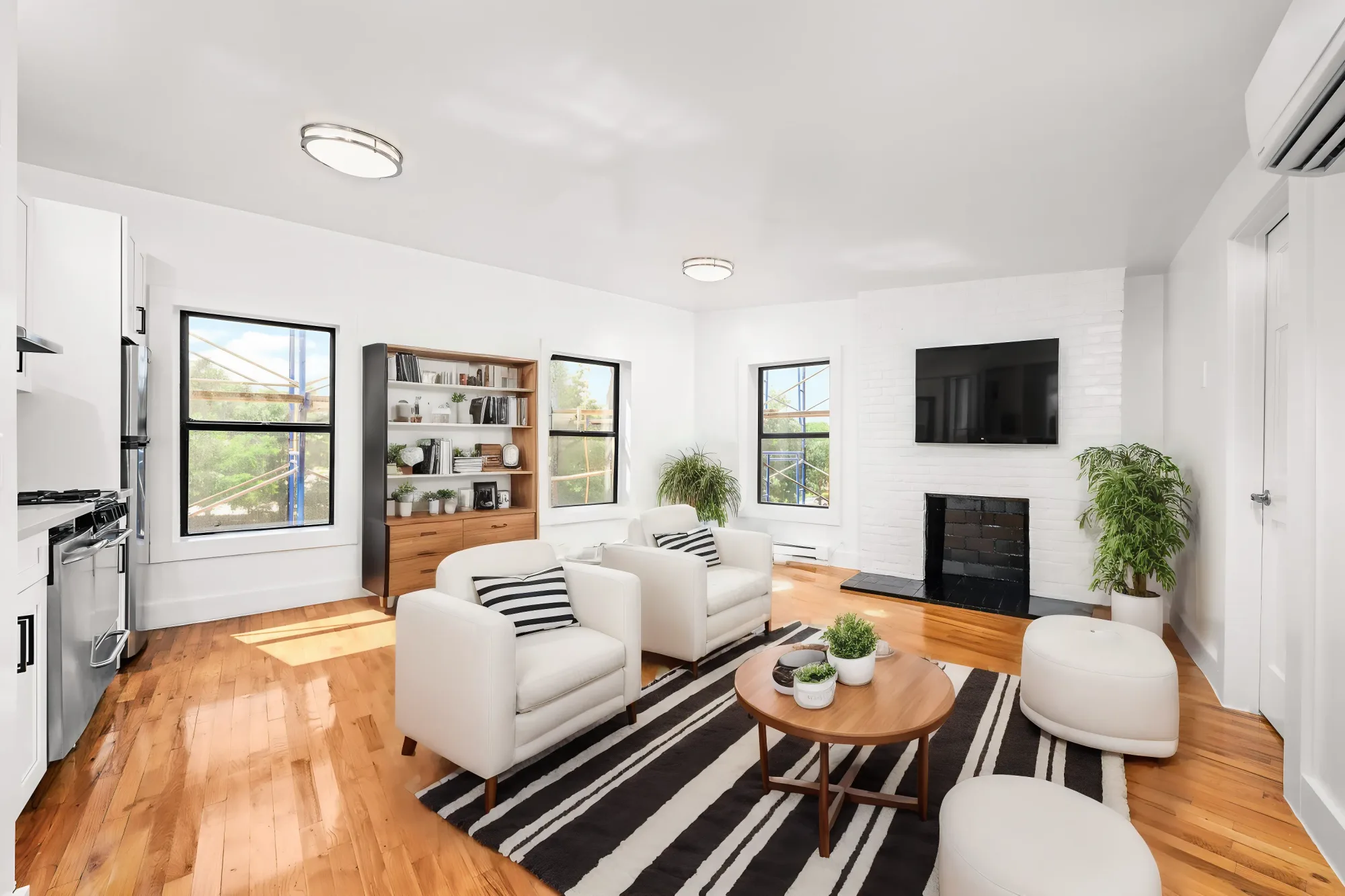 Open-concept living room with hardwood floors, white armchairs, wooden coffee table, fireplace, wall-mounted TV, and multiple windows.