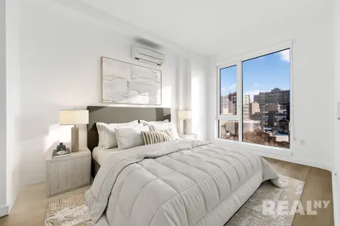 Bedroom with queen bed, white bedding, nightstands with lamps, large windows with city views, and light wood flooring.