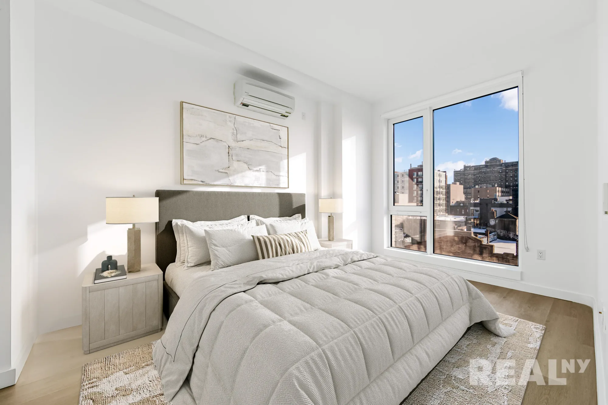 Bedroom with queen bed, white bedding, nightstands with lamps, large windows with city views, and light wood flooring.