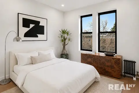 Bedroom with white bed, arc floor lamp, abstract wall art, potted plant, two windows with black frames, and wooden dresser.