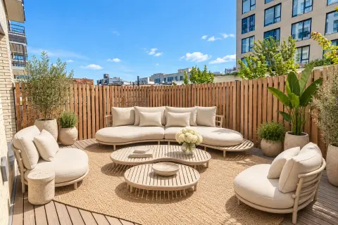Rooftop terrace with wooden fence, beige seating, woven tables, potted plants and city skyline views.