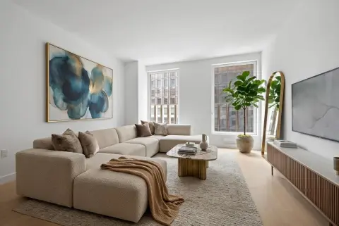 Bright living room with beige sectional sofa, abstract wall art, potted plant, wooden coffee table, and large windows with city views.