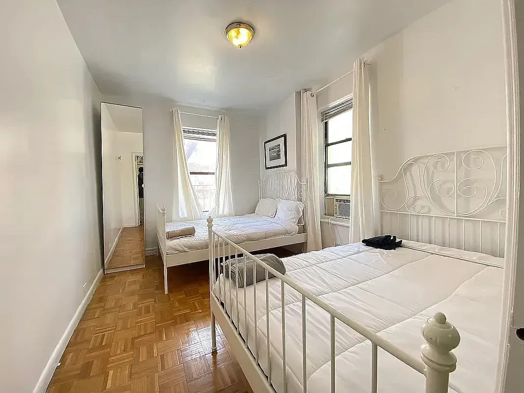 Bedroom with two twin beds with white metal frames, hardwood floors, two windows, and a ceiling light fixture.