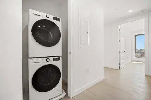 Laundry room with stacked washer and dryer, white walls, light wood flooring, and doorway to hallway.