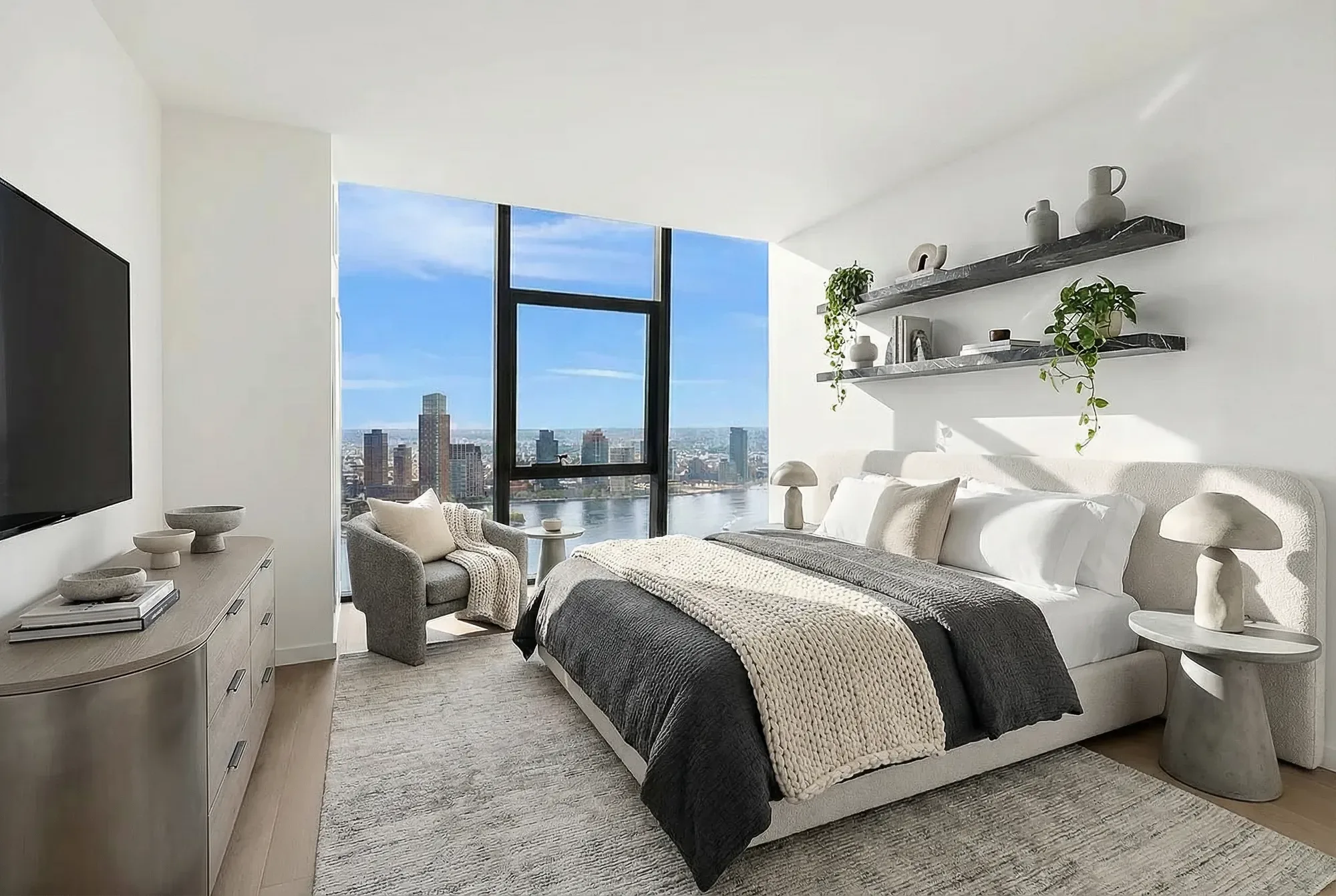 Bedroom with queen bed, floor-to-ceiling windows overlooking city skyline, floating shelves, dresser, and area rug.