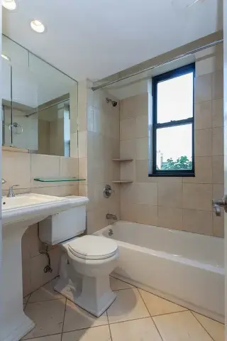 Bathroom with toilet, vanity sink, mirror, bathtub with shower, blue window, and beige tile flooring.