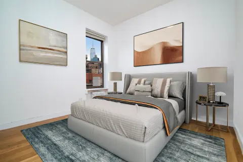 Bedroom with gray upholstered bed, two framed artworks, bedside table with lamp, hardwood floors and blue area rug.