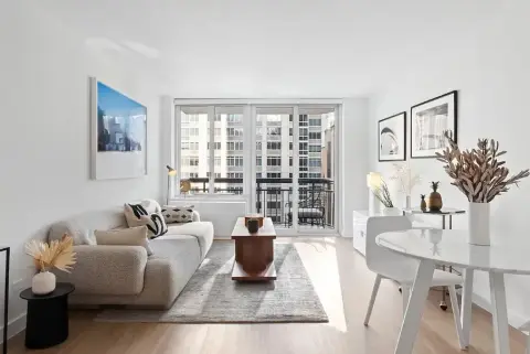 Living room with beige sofa, round white dining table, large windows with city view, and decorative wall art.