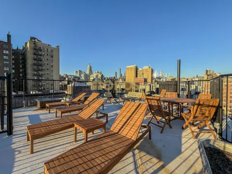 Rooftop terrace with wooden lounge chairs, tables, and city skyline views under clear blue sky.