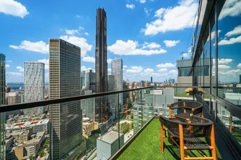 High-rise balcony with artificial turf, wooden chair, and panoramic city skyline views with tall buildings.
