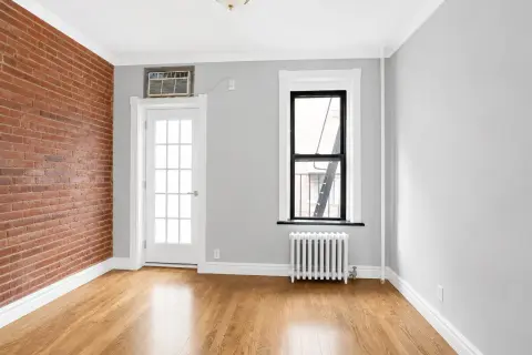 Empty room with exposed brick wall, hardwood floors, two windows, and white radiator.