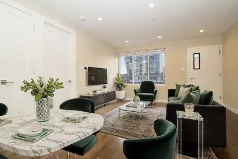 Open-concept living and dining area with marble dining table, black chairs, TV on wall, potted plants, and city views through large windows.