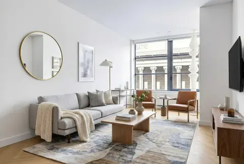 Bright living room with gray sofa, wooden coffee table, round mirror, large windows overlooking a street, and hardwood floors.
