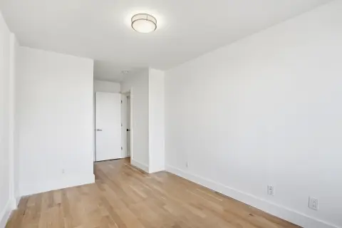 Empty bedroom with white walls, hardwood floors, ceiling light fixture, and doorway to hallway.