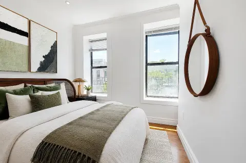 Bedroom with queen bed, green and white bedding, two windows, wooden mirror, framed artwork, and hardwood floors.