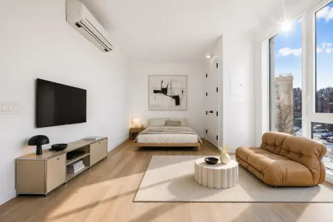 Studio apartment with bed, tan leather chair, TV on wall, wooden furniture, and large windows with city views.