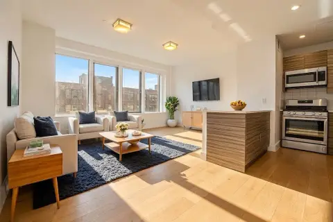 Open-plan living and kitchen area with large windows, hardwood floors, beige sofa, wooden furniture and stainless steel appliances.