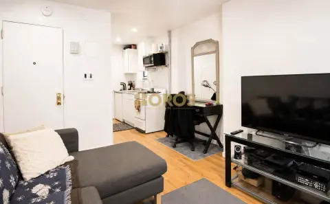 Studio apartment with wood floors, kitchenette, TV stand, mirror, and seating area with ottoman.