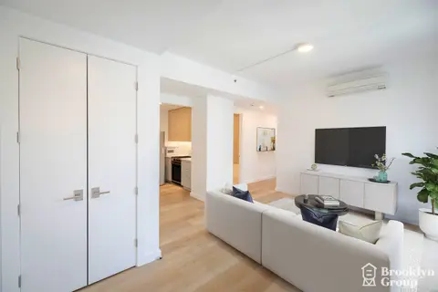 Living room with light wood flooring, white sofa, flat-screen TV on white stand, and white closet doors.