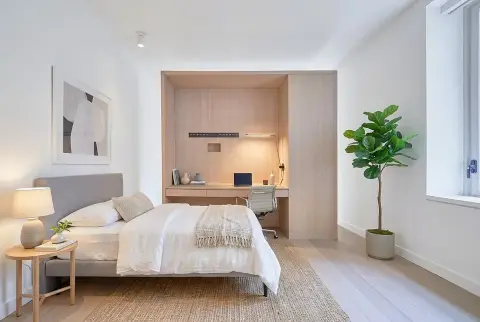 Bedroom with gray upholstered bed, white bedding, wooden nightstand, potted plant, and ensuite workspace visible through open doorway.
