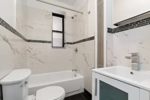 Bathroom with white marble-look tile walls, white vanity with sink, toilet, bathtub, and small window.