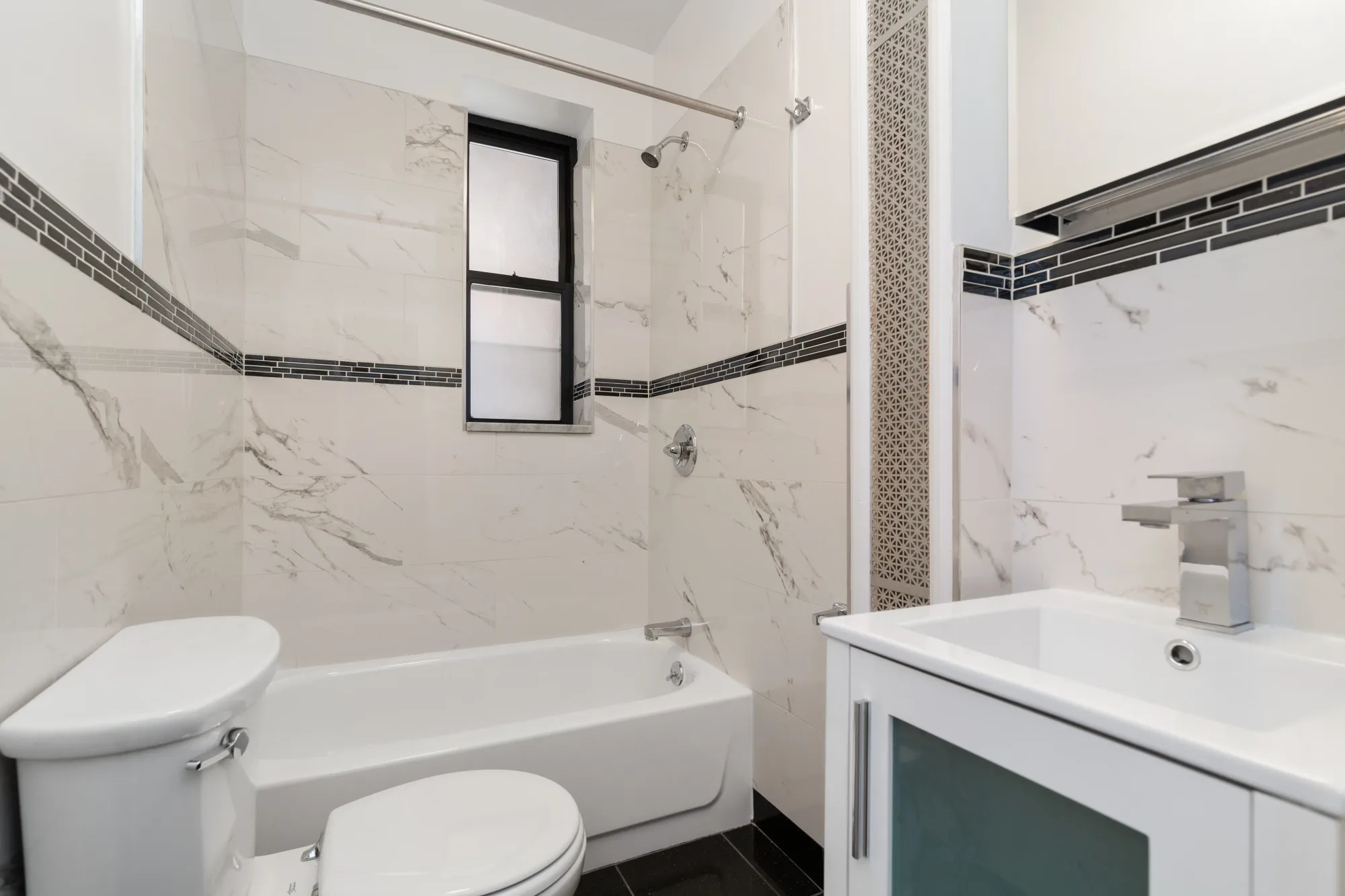 Bathroom with white marble-look tile walls, white vanity with sink, toilet, bathtub, and small window.