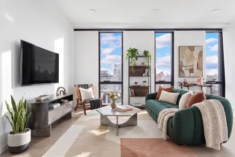 Modern living room with green sofa, wall-mounted TV, floor-to-ceiling windows overlooking city skyline, and light wood flooring.