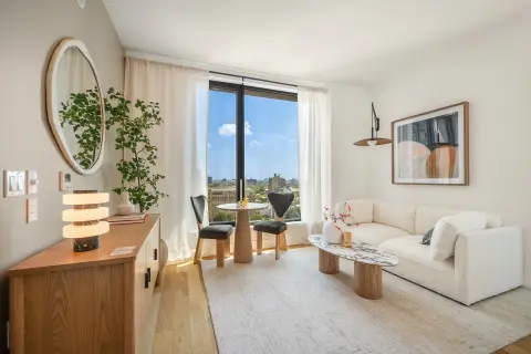 Bright living room with wooden furniture, cream sofa, round mirror, potted plants, and floor-to-ceiling windows with city views.