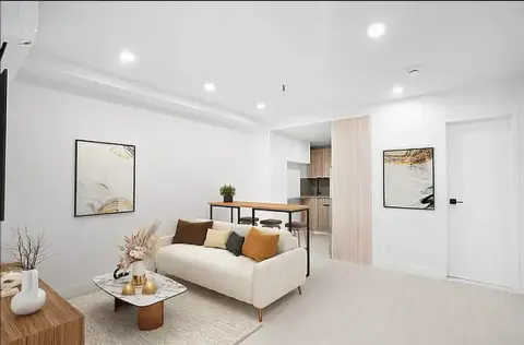 Open-plan living room with cream sofa, wooden coffee table, framed wall art, and kitchen area visible in background.