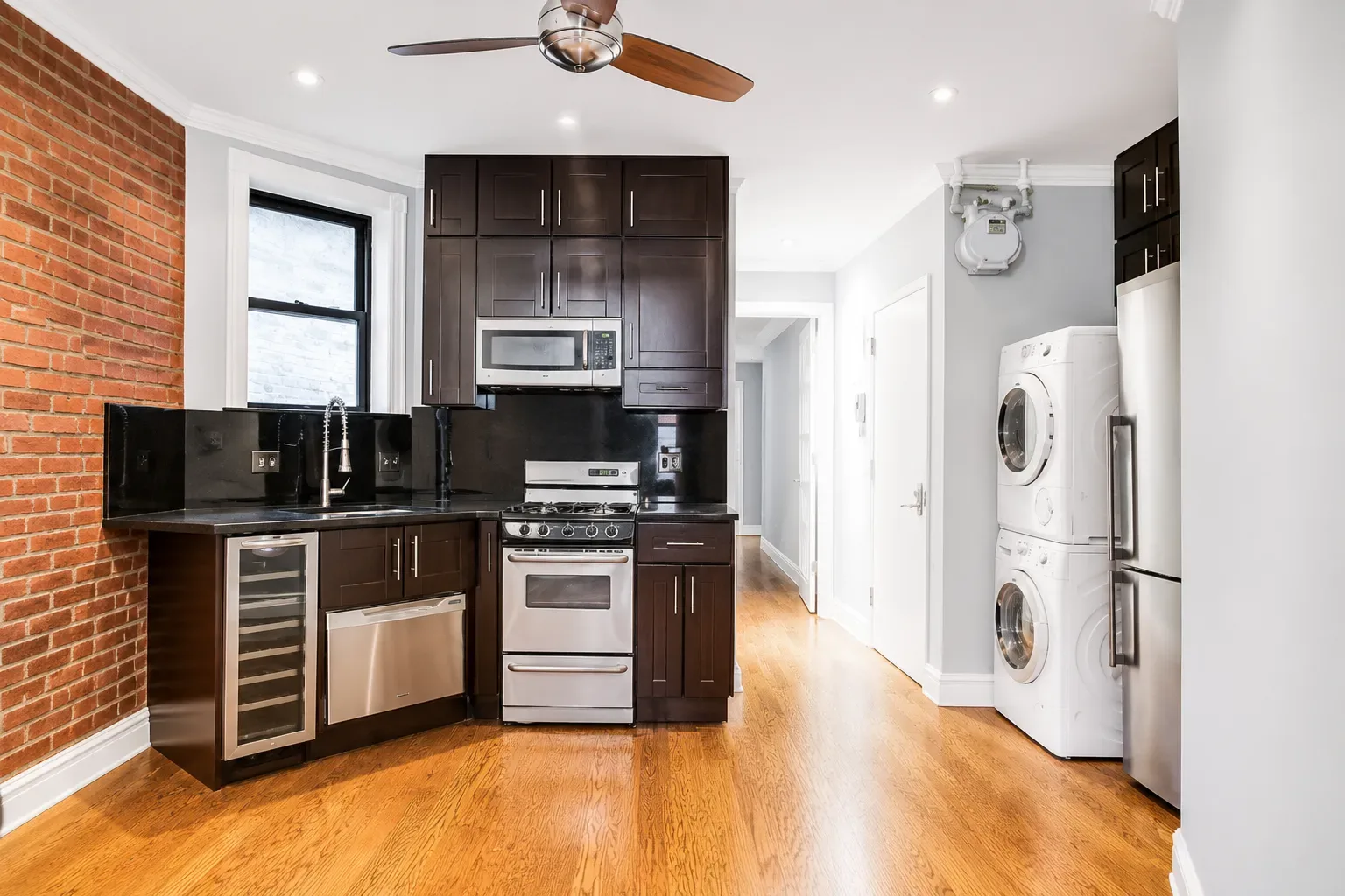 Kitchen with dark wood cabinets, stainless steel appliances, brick accent wall, and stacked washer-dryer on the right.