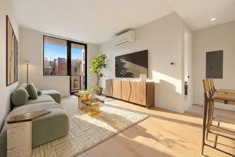 Bright living room with large windows, wooden furniture, green armchair, mounted TV, and city views.