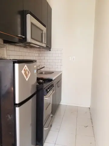 Galley kitchen with stainless steel refrigerator, black gas range, microwave, white subway tile backsplash and light tile flooring.