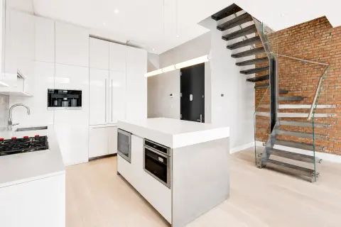 Modern kitchen with white cabinetry, island with built-in appliances, exposed brick wall, and metal staircase.