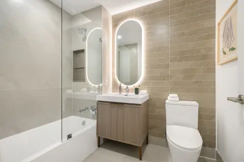 Modern bathroom with dual illuminated mirrors, wood vanity, white toilet, and glass-enclosed shower with tub.