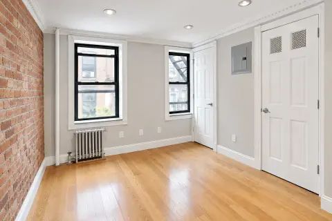 Empty bedroom with exposed brick wall, hardwood floors, two windows, white doors and recessed ceiling lights.