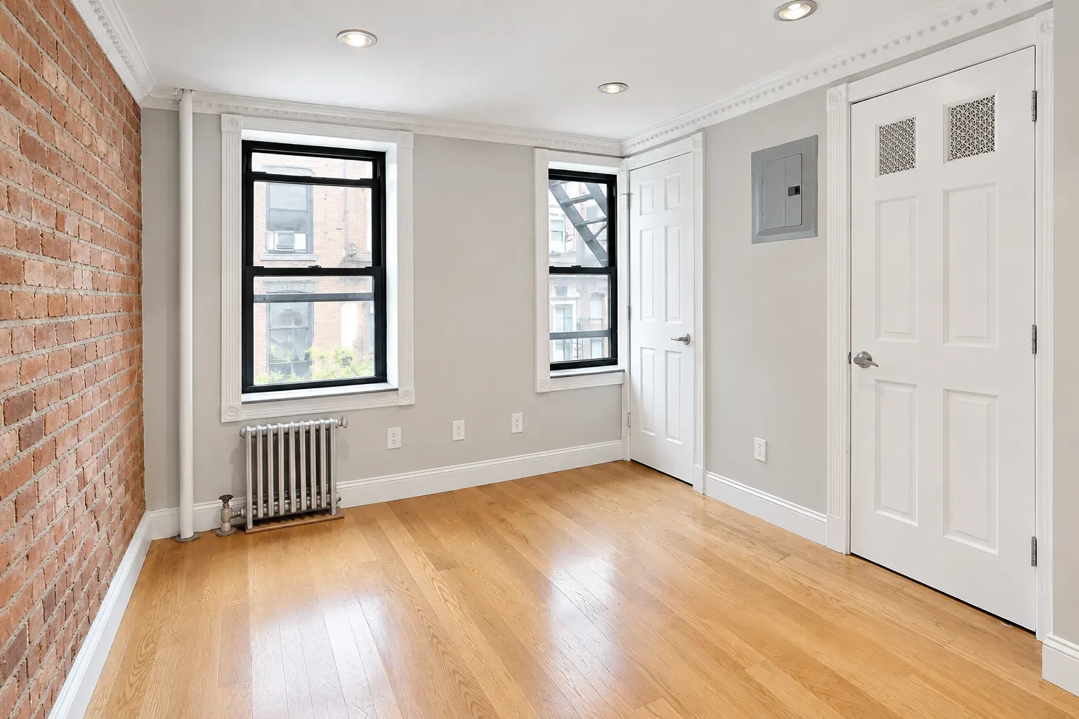 Empty bedroom with exposed brick wall, hardwood floors, two windows, white doors and recessed ceiling lights.